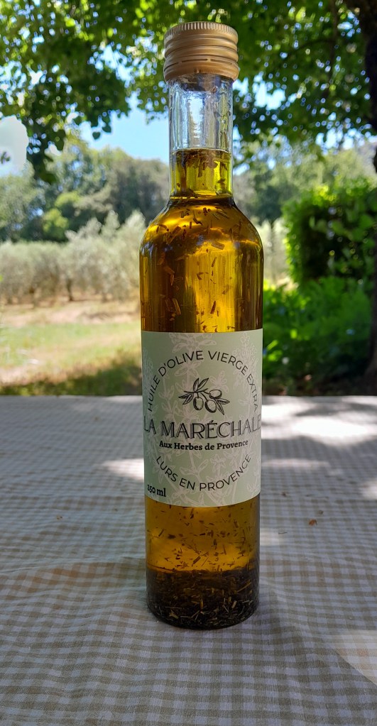 Bouteille en verre contenant de l'huile, sur fonds de vergers d'oliviers. L'étiquette indique : "Huile d'olive vierge extra La Maréchale aux herbes de Provence Lurs en  Provence"