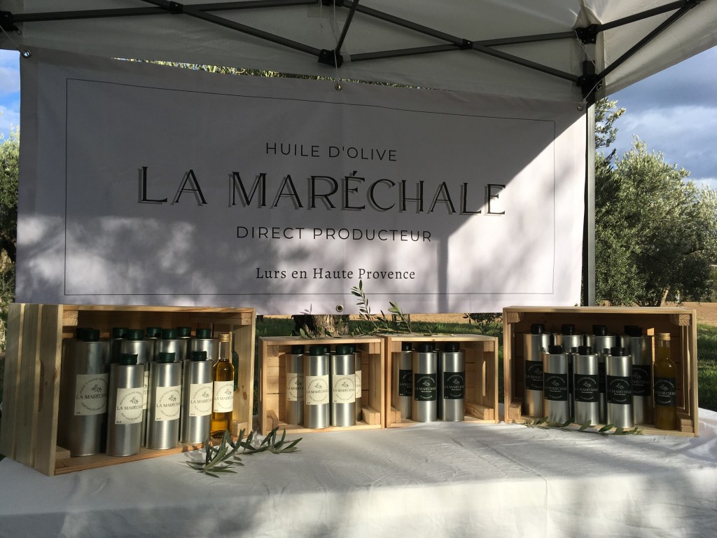 Stand de marché vendant de l'huile d'olive. Une banderole indique "Huile d'olive La Maréchale, direct producteur, Lurs en Haute Provence" Sur la table des caisses en bois contiennent des bidons en inox  remplis d'huile d'olive.